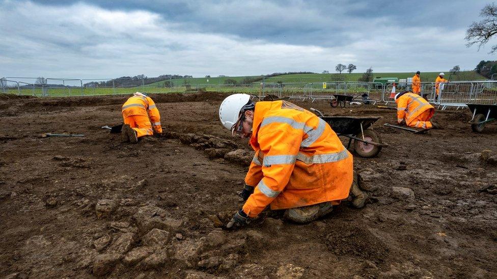 Archaeologists in bright jackets work across a large muddy dig site