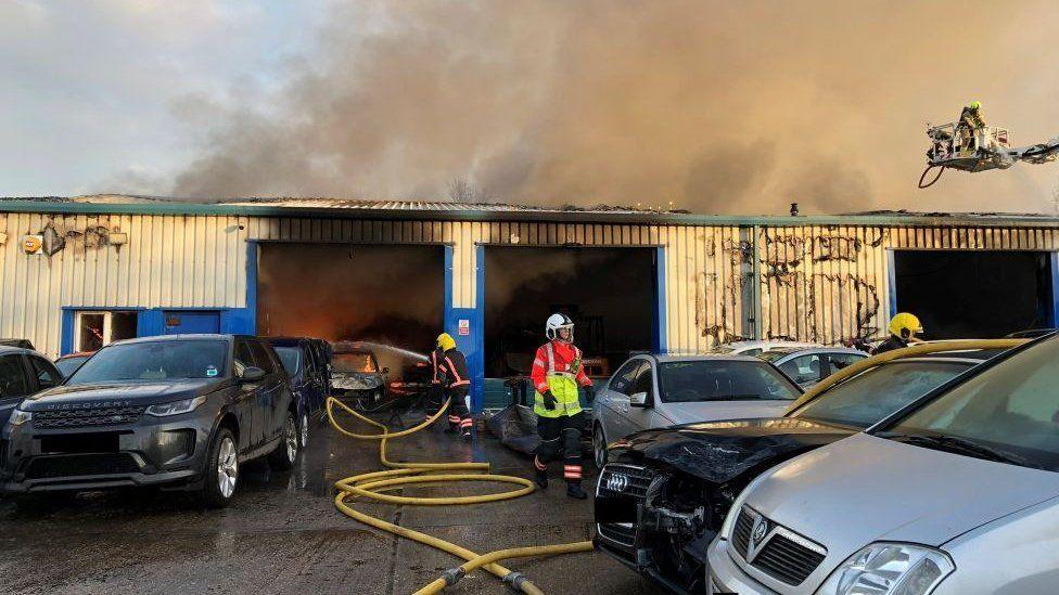 Firefighters are at the scene of a fire at a vehicle recovery yard. There are a number of cars parked outside a large warehouse. Inside, firefighters are putting out a fire in a car. An aerial platform is also being used and there is smoke billowing from the top of the building