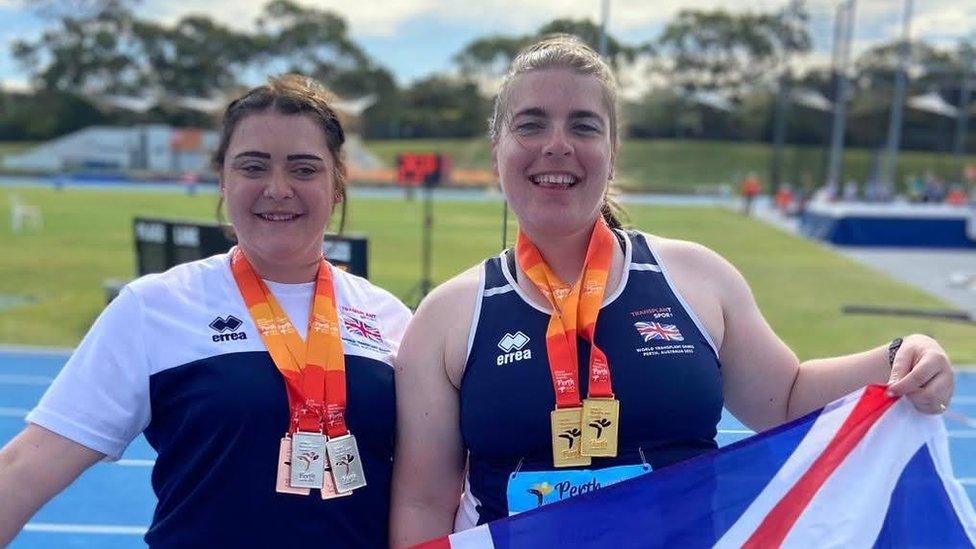 Two young women show off their medals from the British Transplant Games. One is wearing a navy blue running vest, with an "Errea" logo just above her chest on the right and a Union Jack flag opposite on the left. The other wears a white and blue t-shirt with the same decals. They each wear multiple medals around their necks, with thick, bright orange lanyards attached to each one. They're both smiling - it's a joyous scene.