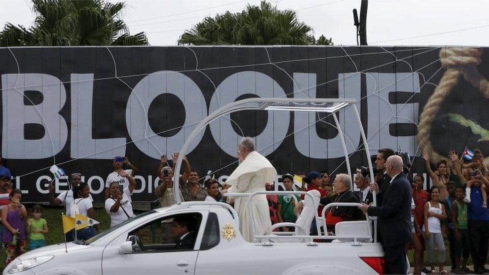 Pope Francis greets Catholic people he passes in front of a billboard that reads "Embargo" outside the airport in Havana on 19 September, 2015