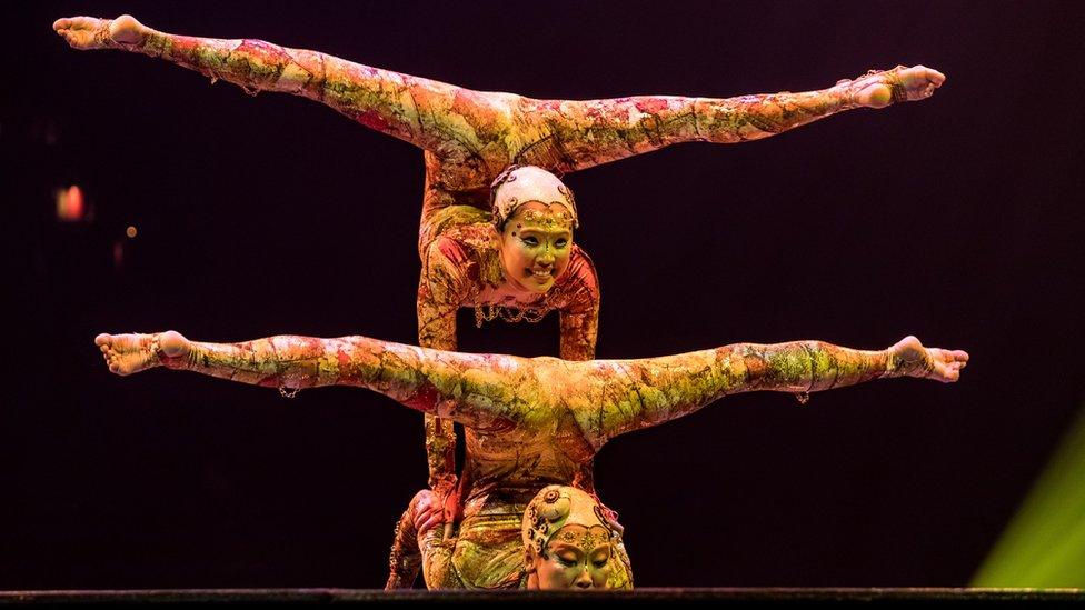 Two Cirque du Soleil acrobats in colourful outfits balance with their legs above their heads