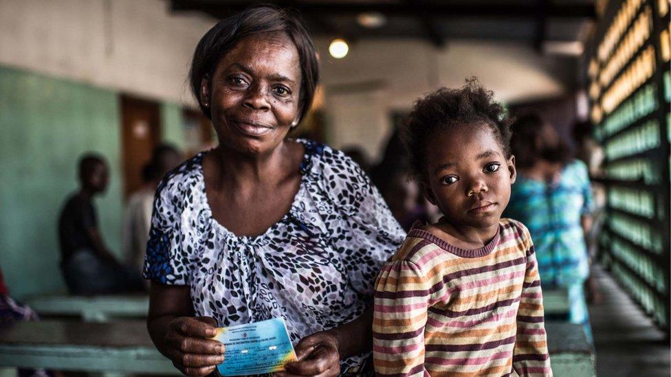 young girl with large dark-brown eyes looks into the camera, with her grandmother sitting next to her