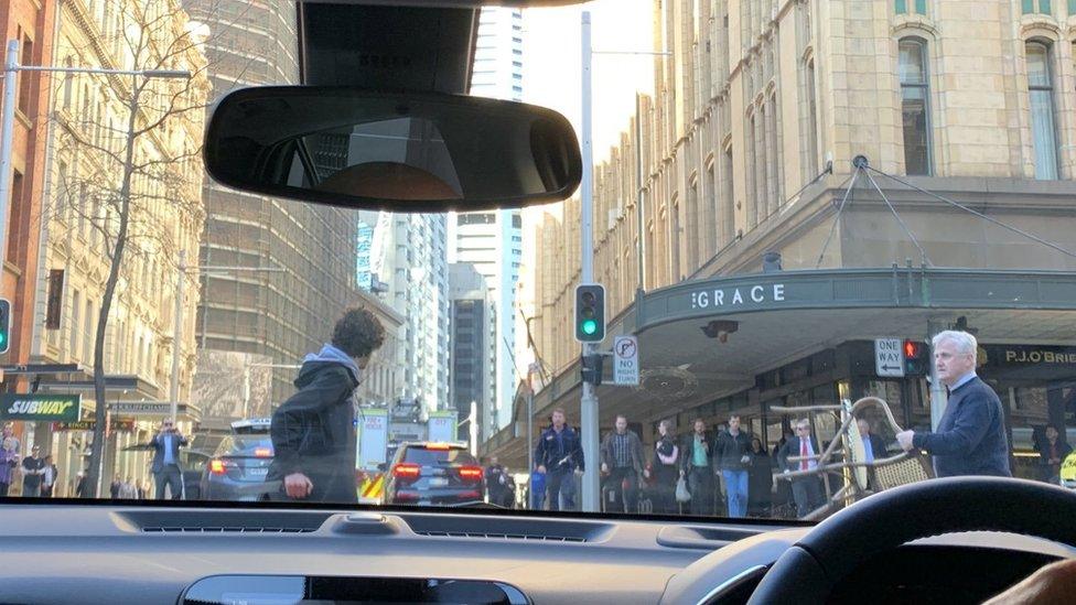 Photo from inside a car shows a man holding a large knife being confronted by a man holding a wicker chair