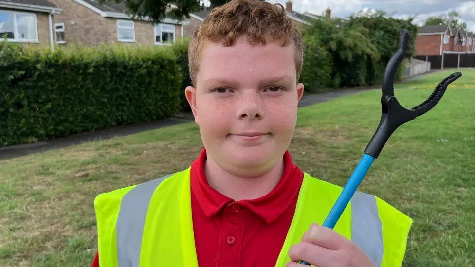 A head and shoulders shot of a boy with short gingery hair, wearing a hi-vis yellow waistcoat over a red polo-shirt. He is holding a blue-handled litter picker up to his shoulder. Behind him is grass, a hedge and some just glimpsed houses. 