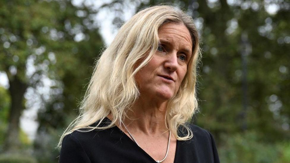 Kim Leadbeater stood in front of green trees, wearing a black top and speaking to a camera.