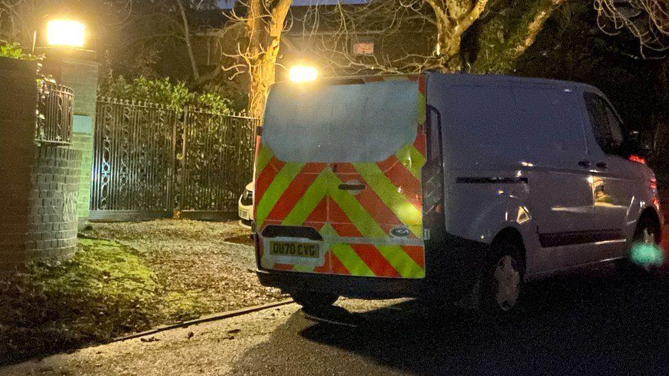 A police van is parked in a residential area, near to some iron gates. It's an alternate angle to the previous picture. The front of the car can be glimpsed behind the van.