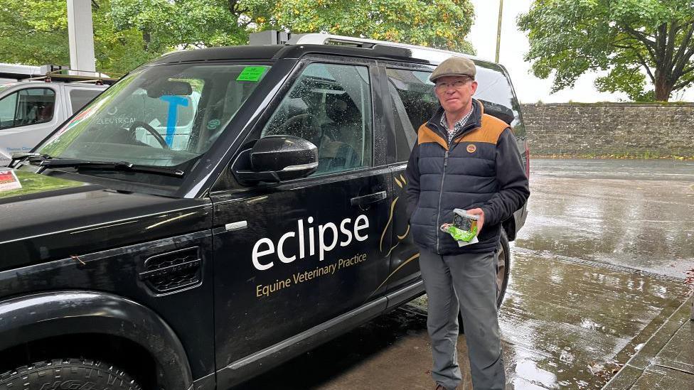 A man wearing a flat cap, and blue and beige bodywarmer over a blue jumper, stands next to a black 4x4 vehicle advertising his equine veterinary practice, on a petrol station forecourt.