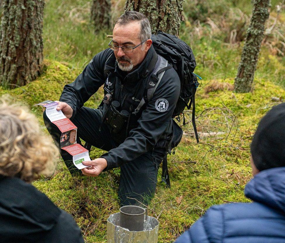 A man in outdoor gear with a backpack and grey hair, a beard and glasses shows a fire advice leaflet to two people - we can only see the backs of their heads. They are outdoors in what looks like a forest and gathered round what could be an unlit camp fire.