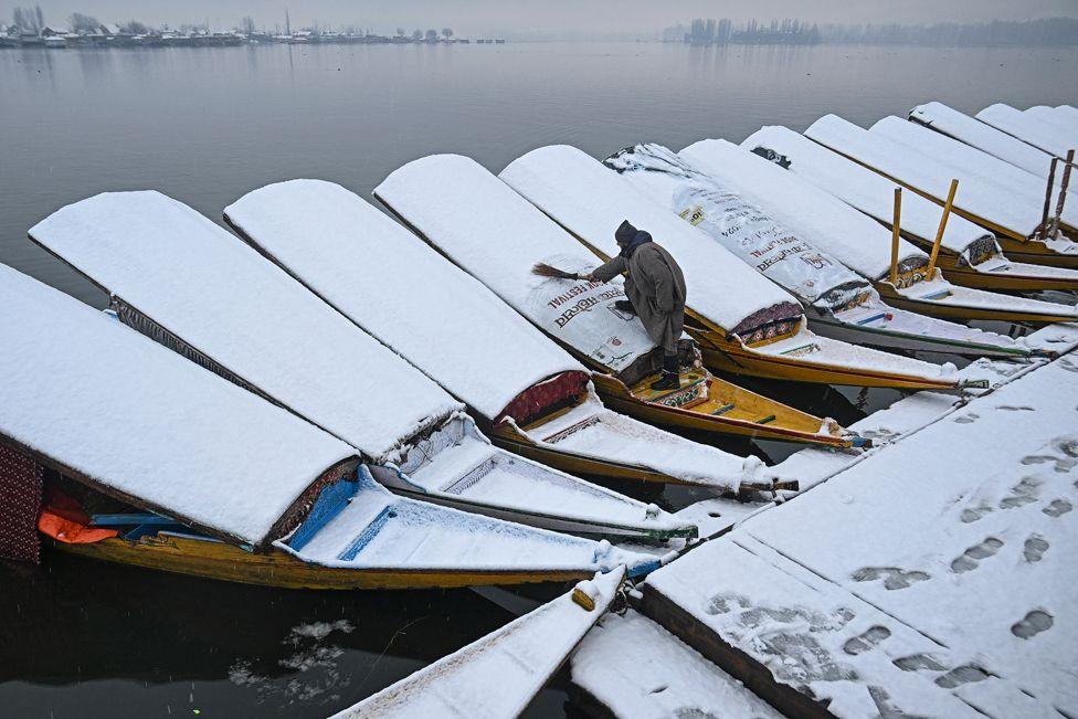 A row of boats covered in snow on a misty lake in India. A man wearing grey uses a brush to remove the snow