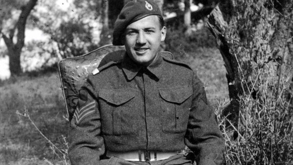 A black and white photograph of Mr Broadhurst in military uniform, when he was younger, wearing a beret. He is sitting down on a hill with trees on it and is looking at the camera.