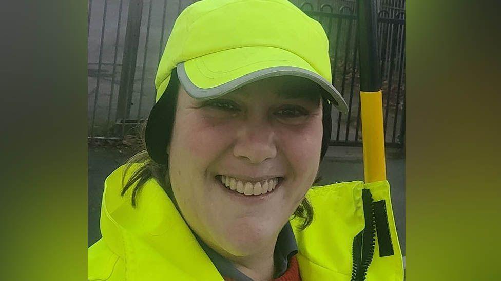Head and shoulders of Rachel Cooper smiling at the camera in a high vis jacket and baseball cap, holding a black and yellow striped lollipop stick