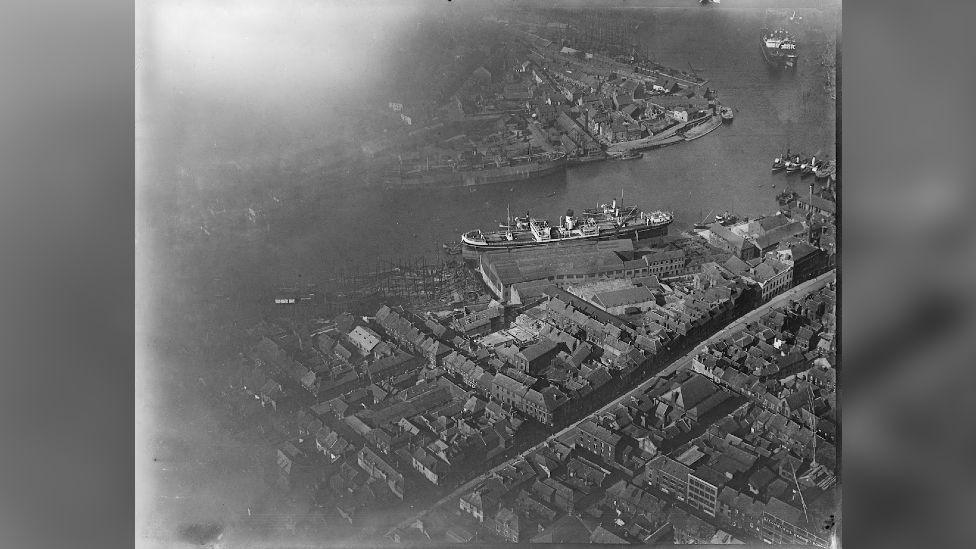 A black-and-white aerial shot of a ship and masses of houses along a large winding river.