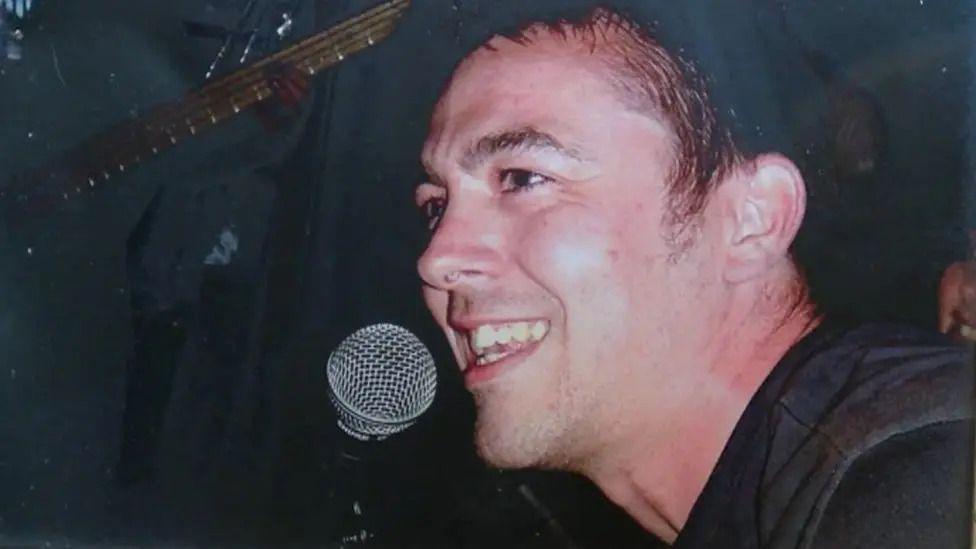 A side profile of a smiling Jon Casey dressed in a black shirt with a silver microphone near his mouth