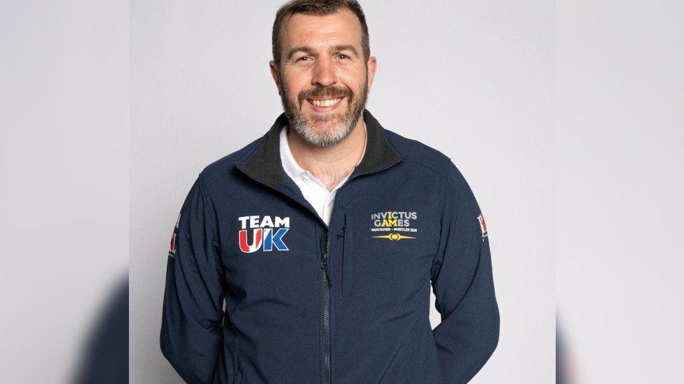 A professional mid-shot of Steve Irwin in a uniform Team UK Invictus Games tracksuit top smiling to camera with a plain white background