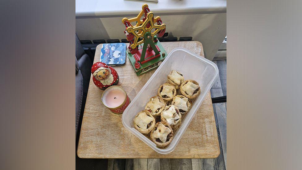 A Tupperware tub filled with mince pies on a table with a Christmas candle and two Christmas ornaments and a bar mat  