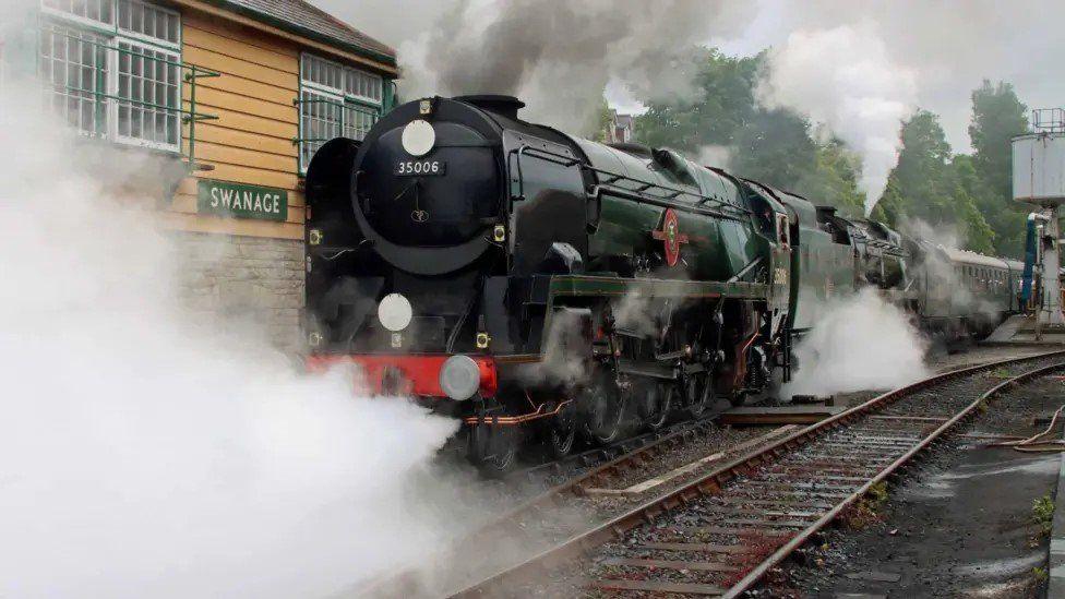 Railway tracks with black and dark green steam train with steam gushing out from underneath outside Swanage railway station