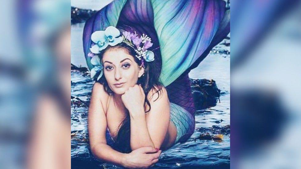 A photo of Ms Seales dressed as a mermaid. She's lying on her front, resting her chin on her hand, and she's wearing a blue and purple mermaid tail and a headdress with blue and purple flowers on. She's got long dark hair.