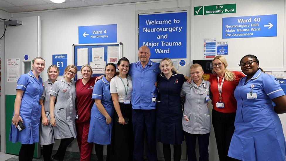 Paul Johnson in blue nurses uniform stands with his arms around the shoulders of his colleagues in front of the entrance to Ward Four at Hull Royal Infirmary