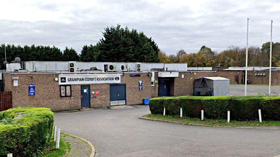 The outside of the Grampian Club, showing a single storey brick building and a driveway to the entrance. To the right are two flag poles.