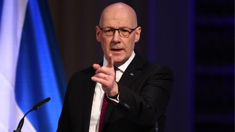 A bald man with glasses, wearing a dark suit and white shirt, points towards the camera while speaking on a stage in front of a dark blue background  