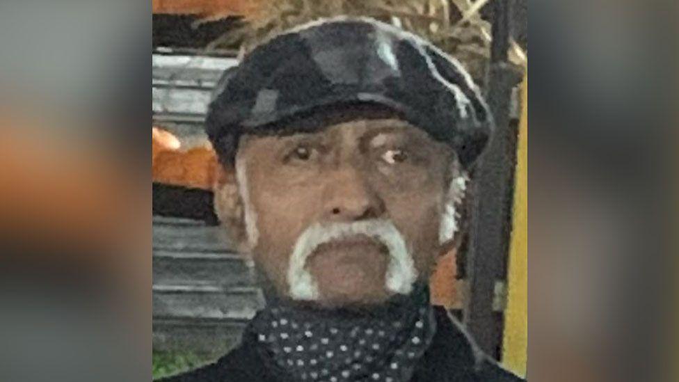 A close up of Chacko Abraham Thenakarayil wearing a black hat and a polka dot neckerchief. He has silver sideburns and a silver handlebar moustache 