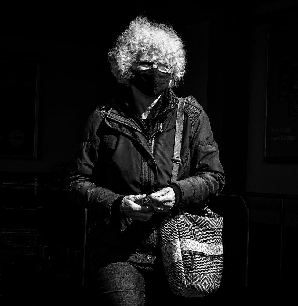 A woman with grey curly hair and glasses walking along wearing a face mask.