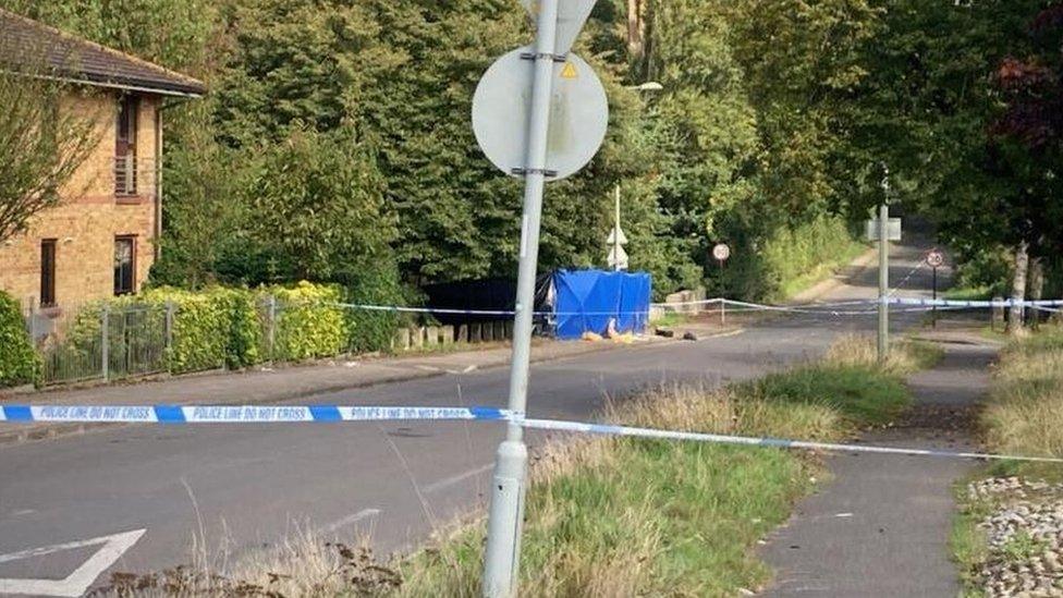 A section of road cordoned off by police tape with a blue tent in the background