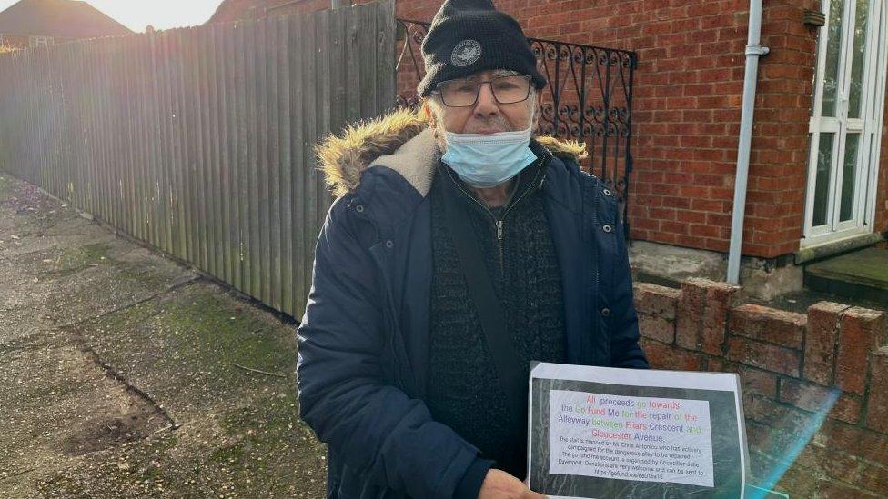 Man wearing blue jacket, mask and woollen hat holds a notice promoting a fundraising campaign