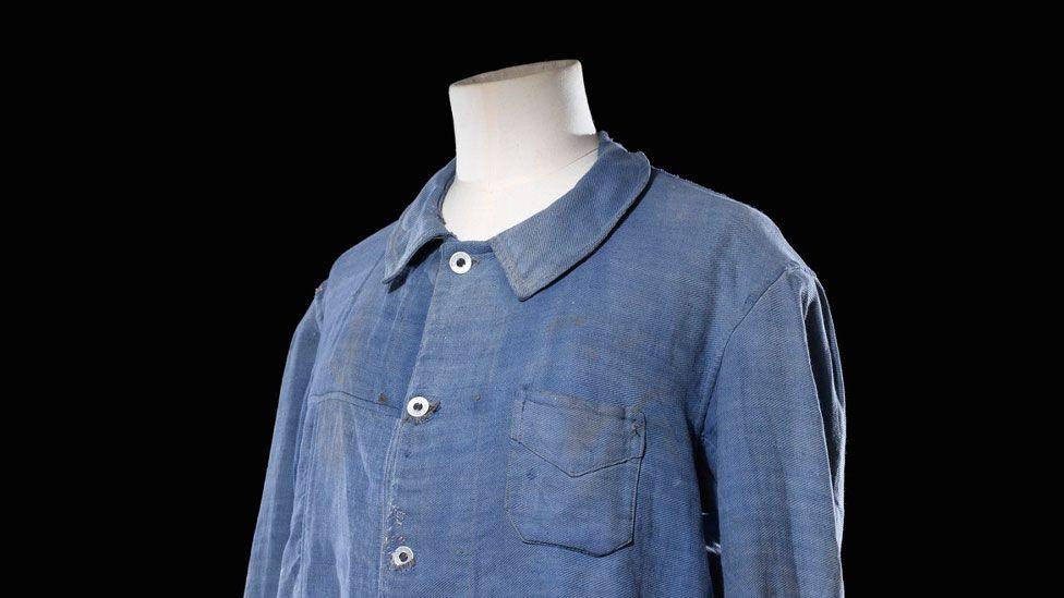 A faded blue heavyweight work shirt with a collar and buttons done up down the front. It has yellow stains on the breast pocket. It is on a headless mannequin. 