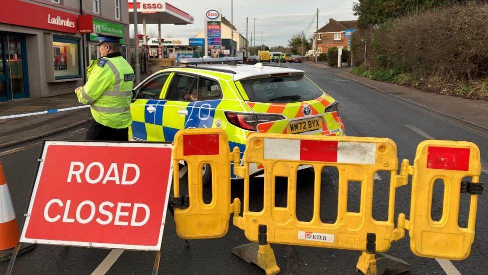 A road closure sign, police car and a police officer standing near some police tape at the foreground of the image. In the background, the A38 stretches out, with an ESSO garage on the left and some more police vehicles in the background.
