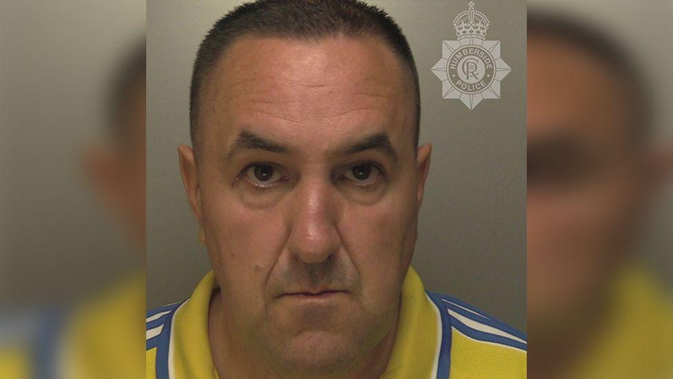 Police custody photograph of Matthew Full he has short black hair and is wearing a yellow shirt with blue and white stripes on the shoulders and collar 