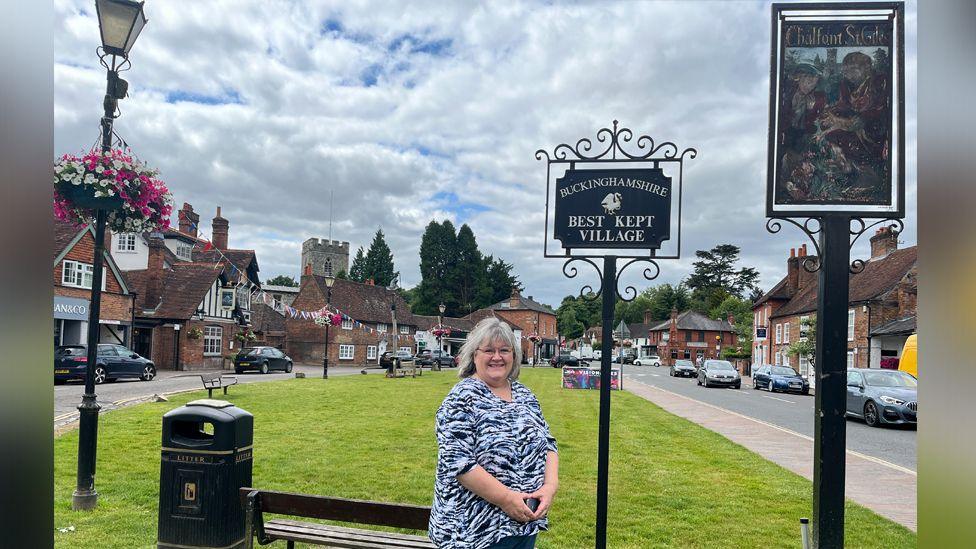 A smiling woman stands on a green with a pretty village scene behind her