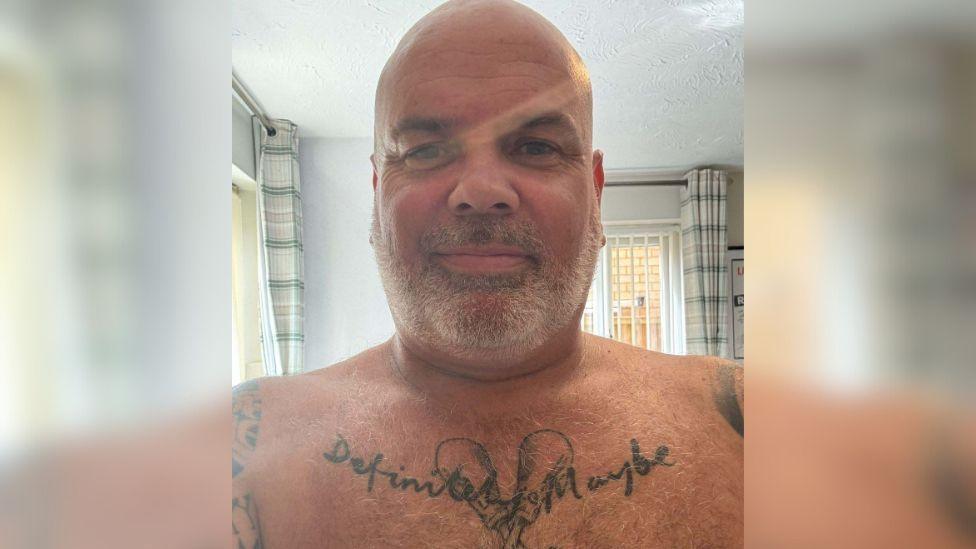 A selfie of John Smallbones from the chest up. He is bald with grey stubble. On his chest is a tattoo saying 'Definitely Maybe' over two maracas.