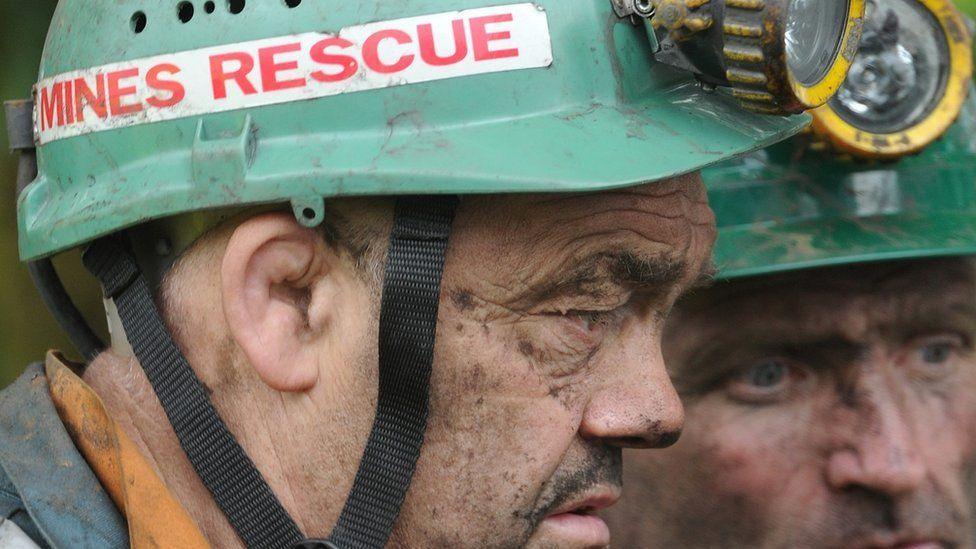 Rescue workers during the Gleision colliery disaster, wearing green miners hard hats with lights on the brong. Two men, one in profile and one face on look sombre. Both have soot on their faces. 