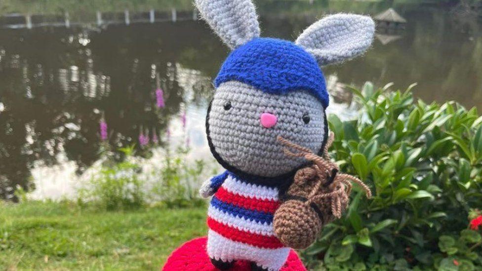 Grey woolly bunny with blue helmet with red, white and blue costume with pond in background