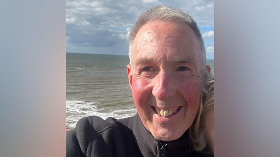 A selfie of Phil Edwards with grey hair and a black top smiling at the camera in front of the sea