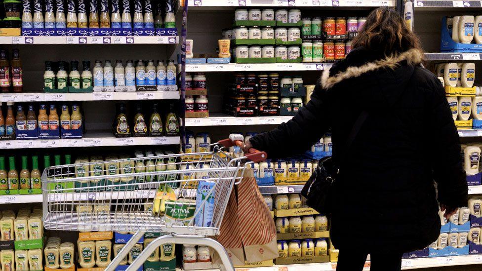 A lady in a supermarket with one hand on her trolley, looking at a shelf filled with condiment bottles.