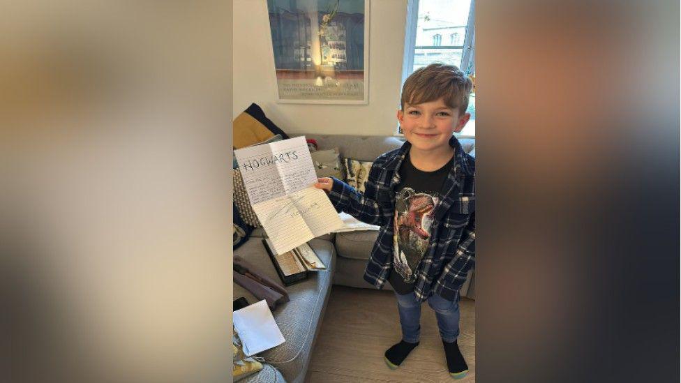 Richard standing and smiling in his family living room and holding the 'Hogwarts' letter sent from Angela Mcgeoch that arrived with his new wand. Richard is wearing a checked shirt over a black dinosaur-print t-shirt, jeans and socks