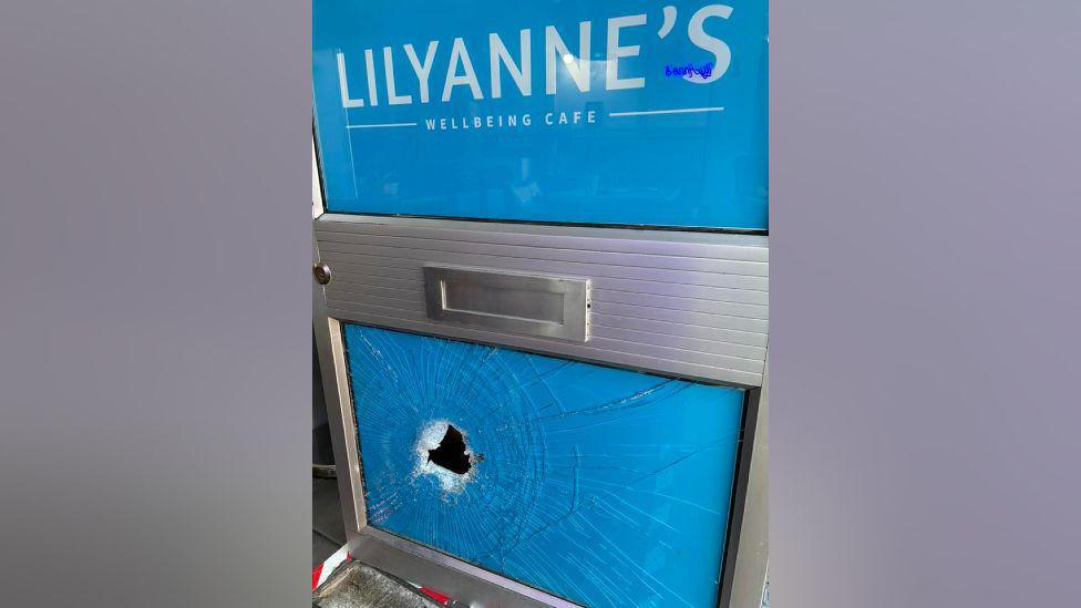 Blue door saying Lilyanne's Wellbeing cafe with a big hole in the glass on the bottom half there are cracks.