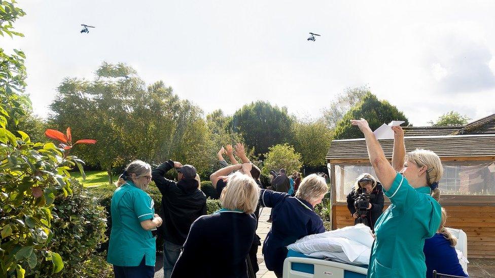 A group of nurses and family around a patient in a hospital bed outdoors, waving and looking up at two small aircraft.