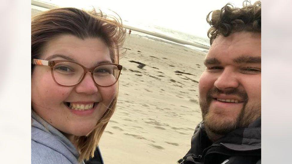 Gabby Evans and Tom Brakewell are smiling in a selfie taken on a beach. Gabby is wearing glasses.