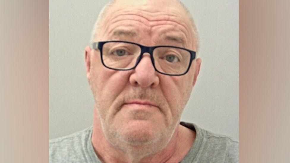 Police custody image of David Berry with receding grey hair and grey beard wearing black glasses and grey t-shirt.