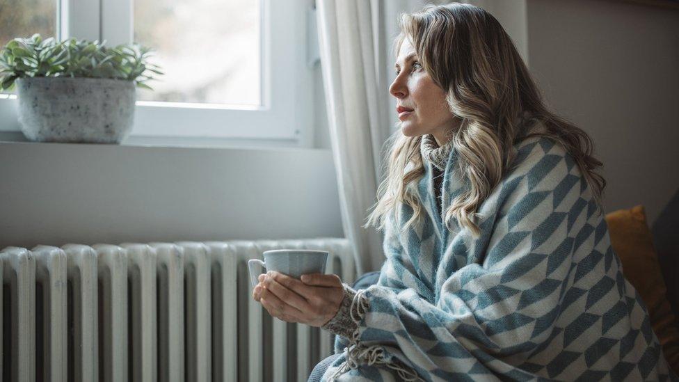 A woman wrapped in a shawl sits next to a radiator