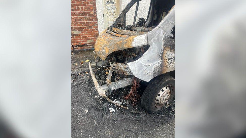A photo of the gutted front of the white van. It is burnt and surrounded by debris. The white door of a bungalow on the street is melted form the fire heat.