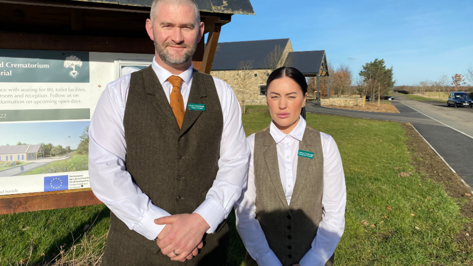 Scott and Laura Clarehugh, are standing in front of a sign saying Northumberland Woodland Burials and Crematorium. They are both young and wearing tweed waistcoats and white shirts. 