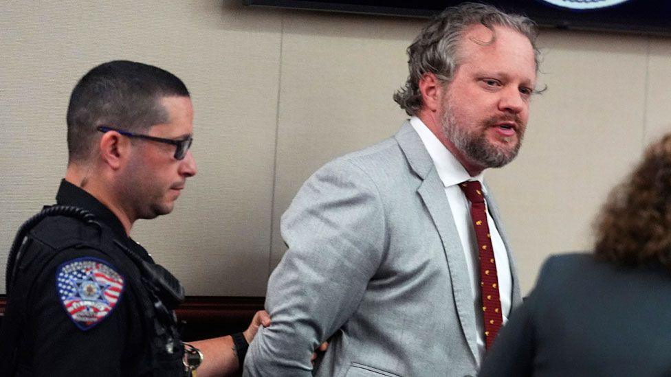 James Craig, wearing a grey suit, appears to be talking to his lawyer as a police officer to the left clutches his arm and leads him away