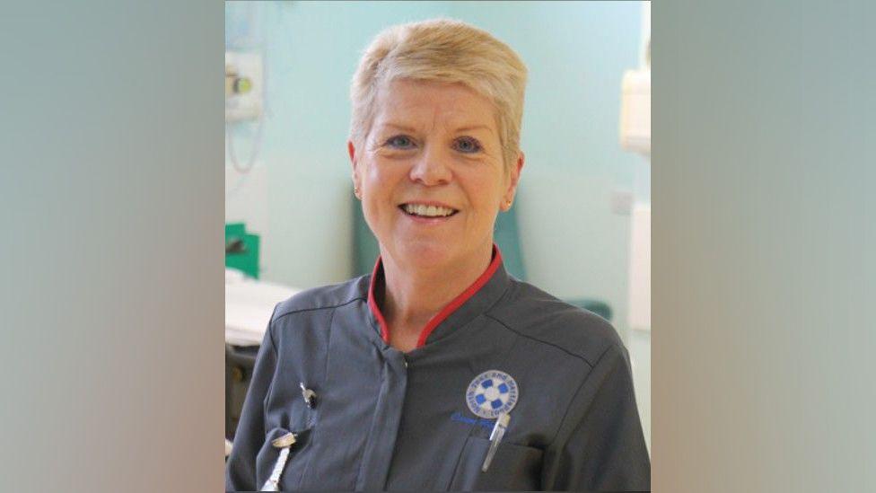 A head and shoulders shot of a nurse with short blonde hair who is smiling at the camera 