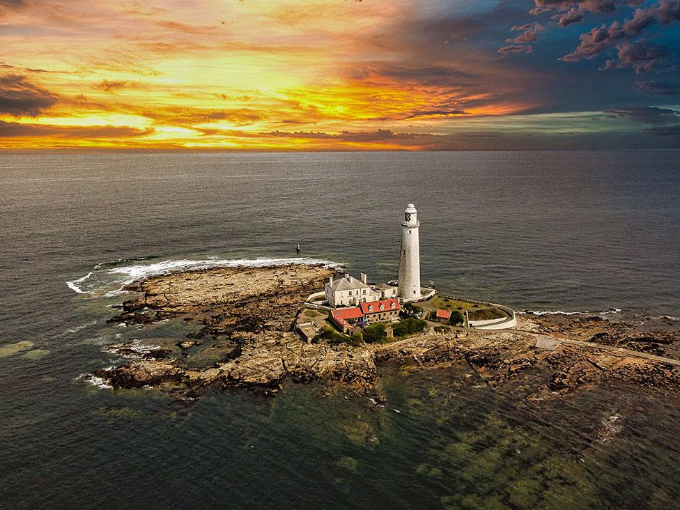 A lighthouse on a rocky outcrop with a sunrise in the background