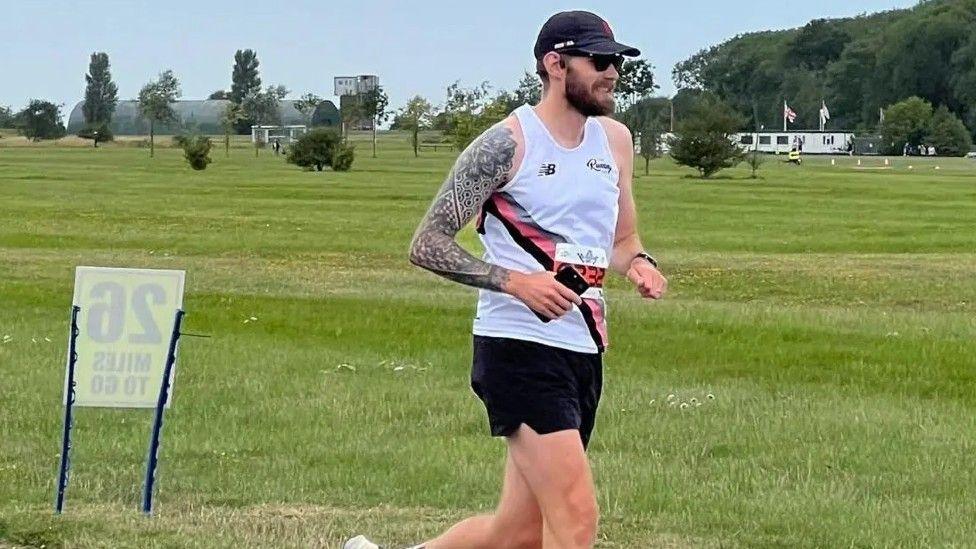 Alex Trivino running wearing a white running vest and black shorts. He has dark brown hair and a beard and is wearing a dark baseball cap.