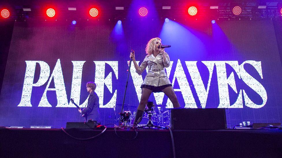 Pale Waves singer, wearing a short black skirt and a silver coat, stands at the front of the stage in front of a guitarist and drummer and a large sign reading Pale Waves at Glastonbury Festival